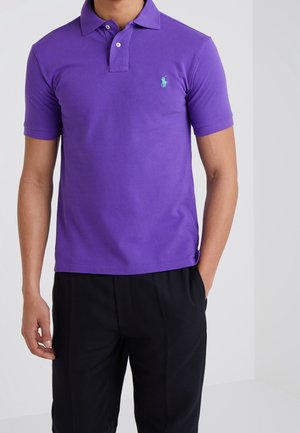 Homme portant un polo violet à manches courtes avec deux boutons et un logo brodé bleu clair, associé à un pantalon noir.