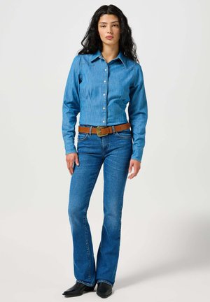 Helleblaue Jeanshemd mit langen Ärmeln und Knopfleiste. Kombiniert mit dunkelblauen Schlagjeans und einem braunen Gürtel mit metallenem Schnallenverschluss. Schwarze Stiefel.