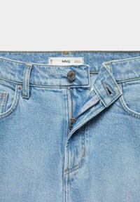 Jean en denim bleu clair avec fermeture éclair partiellement ouverte, bouton en métal fermé et étiquette blanche visible à l'intérieur de la ceinture.