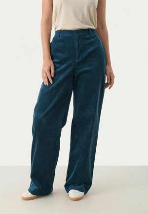 Person trägt hoch taillierte, weit geschnittene Cordhosen in Blaugrün, kombiniert mit beigefarbenem Oberteil und weißen Turnschuhen vor neutralem Hintergrund.