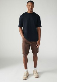 Un tânăr stând, purtând un tricou negru, pantaloni scurți maro, șosete albe și adidași bej, pe un fundal deschis simplu.