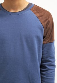 Felpa blu in cotone, con dettagli in suede marrone sulle spalle, collo a costine e maniche lunghe. Texture morbida, vestibilità comoda.