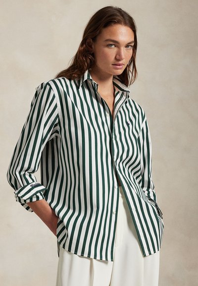 Frau trägt ein grün-weiß gestreiftes Hemd mit Knöpfen, die Ärmel hochgekrempelt, kombiniert mit weißer Hose, steht vor einem neutralen Hintergrund.