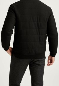 Svart vadderad sweatshirt med en texturerad tyg, ribbade ärmslut och nederkant. Buren över grå herringbone byxor, sedd bakifrån.