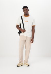 Camiseta blanca con ribete naranja y un diseño gráfico. Pantalones beiges con un ajuste relajado. Zapatillas casuales con una mezcla de acentos en beige y negro.