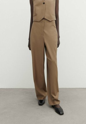 Personne à la peau foncée portant un pantalon taille haute large beige sur mesure, un gilet assorti et des chaussures noires à bout carré, debout sur un sol gris.