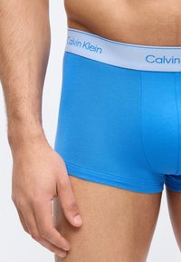 Άνδρας που φοράει φωτεινά μπλε μποξεράκια Calvin Klein, δείχνοντας τον κορμό, το άνω μέρος του μηρού και το αριστερό χέρι που ακουμπάει στο πλάι.