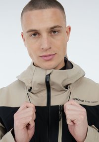 Jonge man die de trekkoorden van een beige en zwarte jas met rits en het "PROTEST PVRE GREEN" logo op de borst aanpast tegen een effen achtergrond.