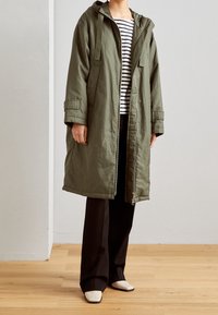 Parka verde oliva con cappuccio, chiusura con zip e tasche laterali; abbinata a una maglietta a righe e pantaloni neri, in piedi su un pavimento di legno.
