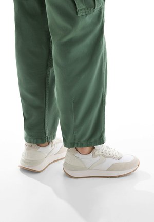 Baskets blanches avec des accents beige clair, des panneaux texturés et des semelles en caoutchouc, associées à un pantalon vert en tissu léger et ample.