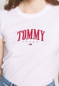 White cotton t-shirt with a round neckline. Features bold red text "TOMMY" and "JEANS" below, with a small logo in red, white, and blue.