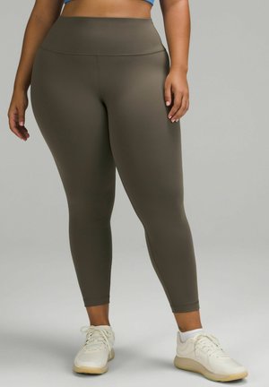 Olive green leggings made of stretchy fabric with a high waistband, ankle-length cut, and smooth texture. White athletic shoes complete the look.