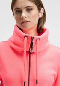 Pink zip-up hoodie with high neck, featuring a textured fabric and contrasting zipper details. Drawstrings are thick and textured.