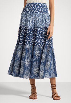 Kvinde iført en blå lagdelt midi nederdel med hvide blomsterprint og brune flade sandaler med remme, står foran en ensfarvet baggrund.