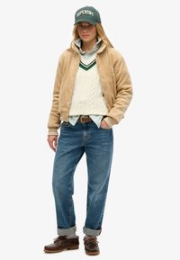 Tan cordjack, crèmekleurige kabelgebreide trui, groen gestreept shirt, blauwe jeans en donkerbruine schoenen. Groene baseballpet met het "SUPERDRY" logo.