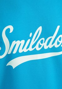 Camiseta turquesa con un gran texto en cursiva blanca "Smilodon" y un elegante trazo decorativo debajo, textura suave y fabricada en material sintético.