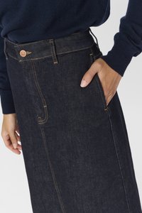 Main de femme dans la poche d'une jupe en denim foncé avec surpiqûres contrastantes, portant un haut à manches longues bleu marine sur un fond clair.