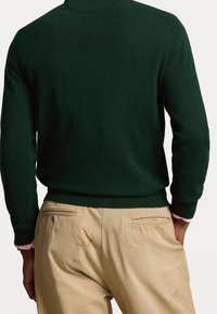 Man draagt een donkergroene trui met lange mouwen en beige broek, handen gedeeltelijk in de broekzakken, gezien van achteren tegen een witte achtergrond.