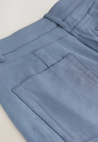Hellblaue, weiche Hose mit einer hinteren Tasche und Gürtelschlaufen; entlang des Taillenbunds und der Taschenkanten ist eine detaillierte Naht sichtbar.