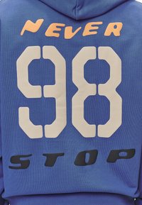 Felpa blu con grande numero bianco "98" e testo arancione "NEVER" sopra, testo nero "STOP" sotto, realizzata in tessuto testurizzato.