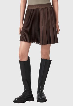 Femme portant une jupe plissée marron associée à des bottes en cuir noires mi-hautes sur un fond clair.