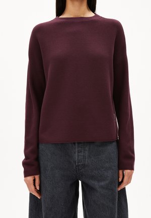 Femme portant un pull à manches longues, col rond, de couleur violet foncé, et un jean en denim noir ample, debout devant un fond uni.