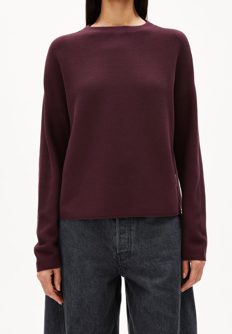 Femme portant un pull à manches longues, col rond, de couleur violet foncé, et un jean en denim noir ample, debout devant un fond uni.
