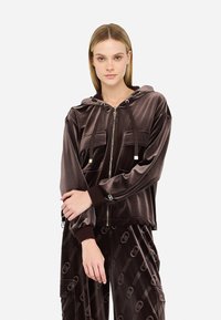 Chaqueta de terciopelo con capucha en marrón oscuro, con dos bolsillos frontales, cierre de cremallera y puños y dobladillo acanalados. Acentuada con detalle de logo.