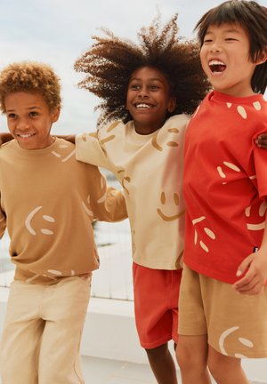 Tre bambini sono con il braccio attorno alle spalle, indossando abbigliamento casual: magliette beige e rosse con motivi giocosi e pantaloni corti nei colori coordinati.