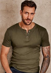 Olive green short-sleeve henley shirt with three-button placket. Soft fabric, fitted design, features visible tattoos on the arm.