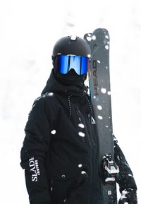 Musta hiihtopuku vetoketjullisella hupulla, musta kypärä, siniset peililasit ja harmaat sukset, joissa on musta siteet. Ilmassa näkyy lumihiutaleita.