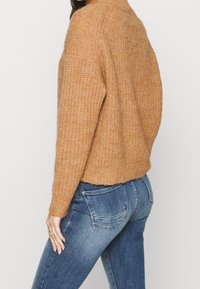 Pull marron en tricot avec un col montant et une texture côtelée, associé à un jean en denim bleu avec des poches arrière.