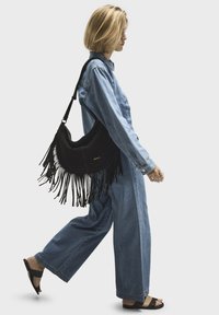 Black suede shoulder bag with fringes, featuring a curved design. Model wears denim outfit and flat sandals, walking in a minimalistic setting.