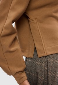 Hanorac maro cu o croială lejeră, manșete cu nervuri și detalii de cusătură la margine, asortat cu pantaloni cu model cu carouri.