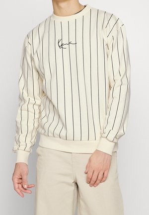 Mężczyzna ubrany w kremową koszulę z długim rękawem z cienkimi pionowymi czarnymi paskami i kursywnym logo na piersi, w połączeniu z jasnobeżowymi spodniami.