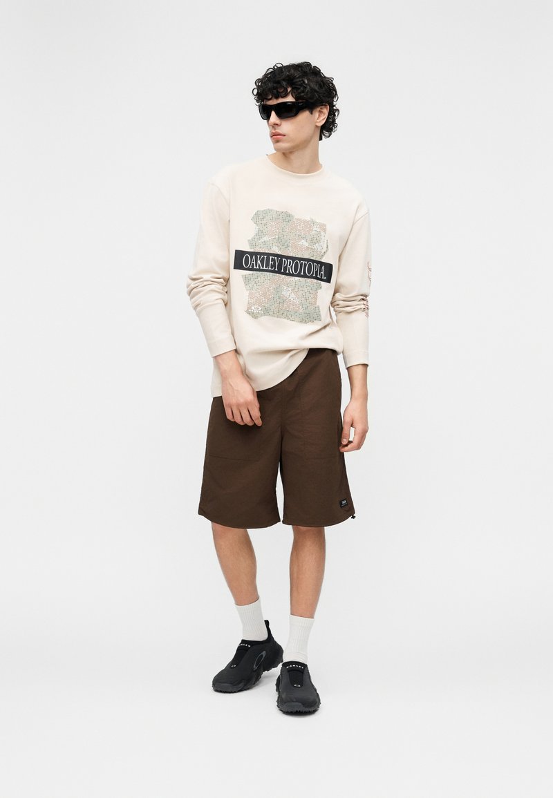 Young man wearing beige Oakley Protopia sweatshirt, brown knee-length shorts, black sneakers, white socks, and black sunglasses, standing against white background.