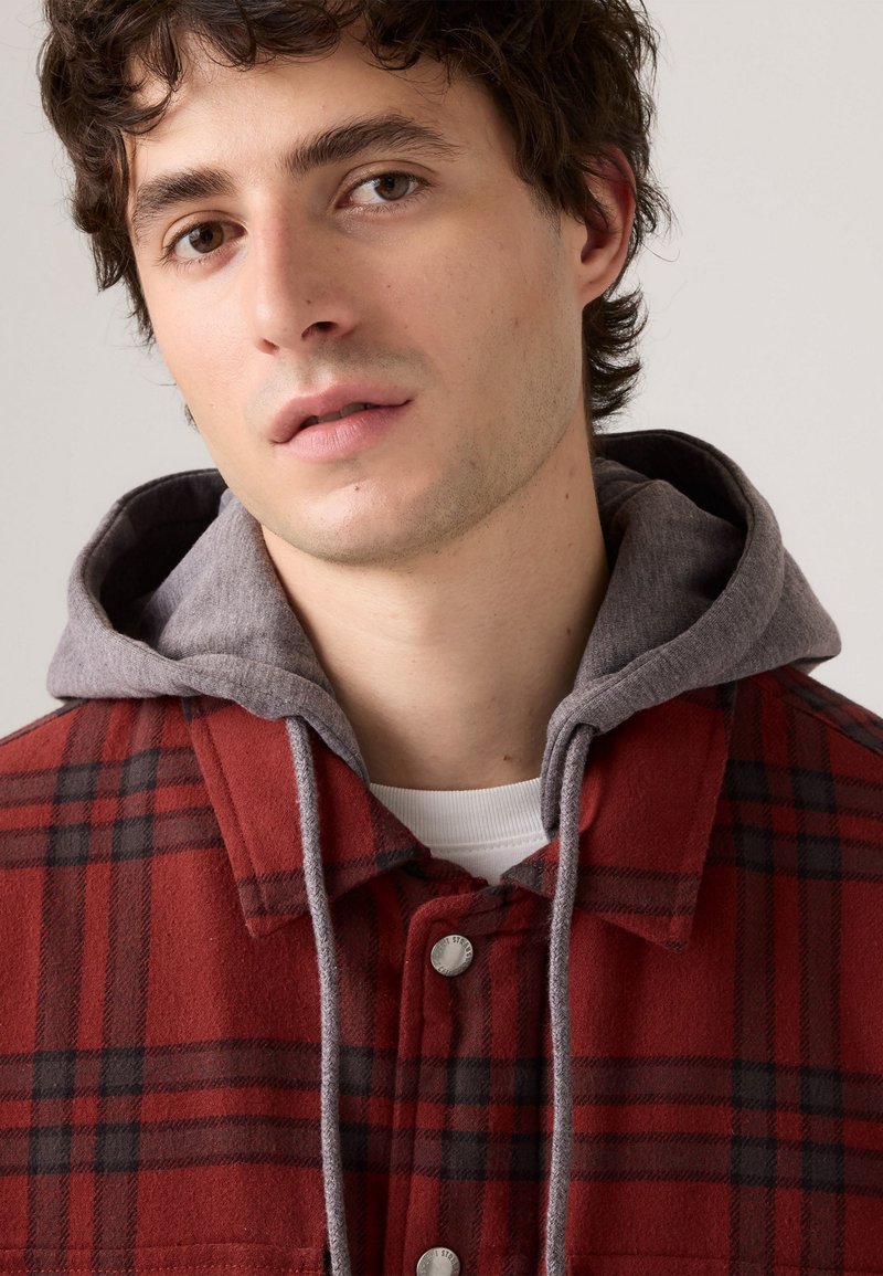 Czerwono-czarna kurtka w kratę z szarym kapturem, z zapięciem na guziki, teksturowanym materiałem i białą koszulą pod spodem.