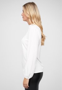Maglietta bianca a maniche lunghe con una vestibilità comoda e scollatura rotonda. Le maniche presentano polsini elasticizzati. Abbinata a pantaloni neri attillati.