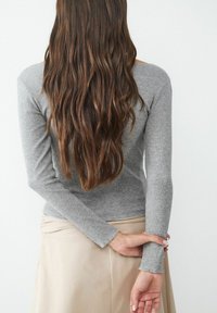 Gray long-sleeve ribbed top with fitted sleeves, paired with a light beige skirt. Hair is long and wavy, visible from the back.