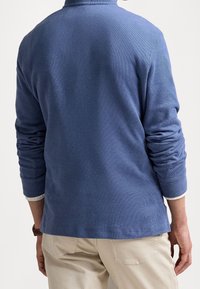 Pulóver texturizado de color azul claro con mangas largas y cuello alto. El dobladillo es ligeramente curvado y una capa interior blanca es visible en los puños.