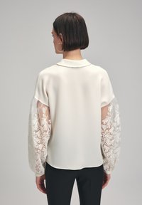 White blouse with sheer, embroidered sleeves featuring floral patterns. The design includes a classic collar and a smooth, opaque body.