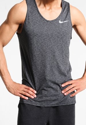 Mężczyzna w ciemnoszarej sportowej koszulce na ramiączkach z logo Nike, dłonie opierające się na biodrach na jednolitym jasnym tle.