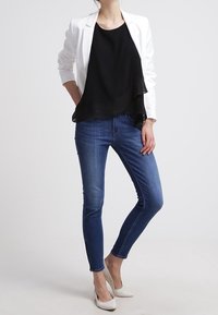 Blazer bianco su un top nero a strati, abbinato a jeans blu aderenti e ballerine bianche, che mostrano un design minimalista e moderno.