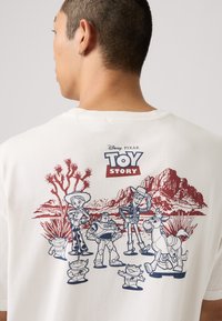 Bílý bavlněný tričko s potiskem červených a námořnických postav z Toy Story na pozadí pouštní krajiny. Krátké rukávy a design s kulatým výstřihem.
