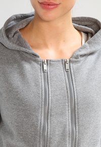 Felpa grigia con zip, dotata di doppi zip in metallo, texture morbida e cappuccio. Il tessuto appare caldo e casual. Il design include un colletto alto e una vestibilità streamlined.