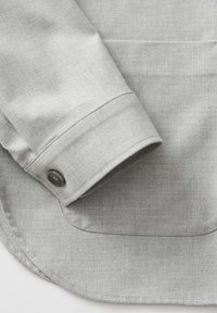 Chemise gris clair en tissu texturé avec un poignet replié et un bouton foncé, mettant en avant un design simple et classique.