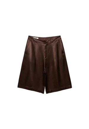 Femme portant un débardeur blanc, un short long marron, une veste marron assortie sur une épaule, des chaussettes grises et des talons hauts marron, debout contre un mur.