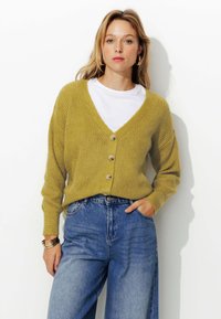 Cardigan tricoté jaune moutarde avec un col en V, doté de cinq boutons et d'une texture côtelée ; associé à un jean bleu taille haute.