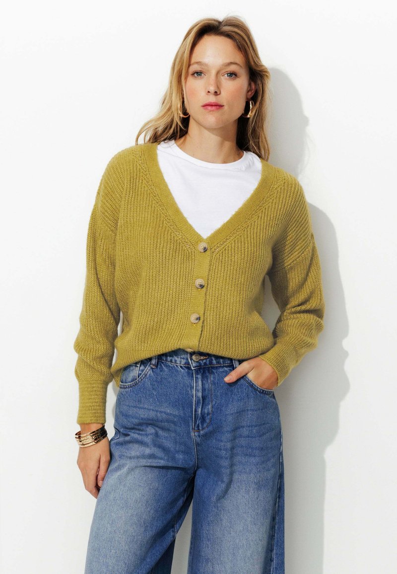 Cardigan tricoté jaune moutarde avec un col en V, doté de cinq boutons et d'une texture côtelée ; associé à un jean bleu taille haute.
