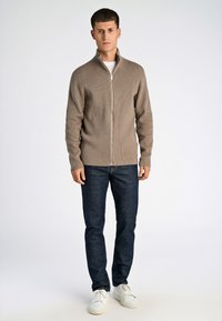 Maglione beige a coste con zip e colletto alto, indossato sopra una t-shirt bianca, abbinato a jeans blu scuro e sneaker bianche.